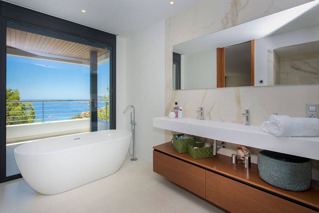 Diseño de cuarto de baño de lujo con una bañera exenta blanca junto a un gran ventanal con vistas al mar