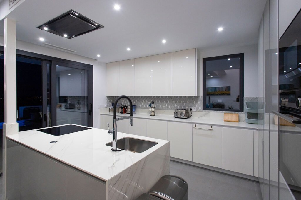 Perspectiva de cocina moderna con armarios en bloque color gris, isla central equipada con placa de cocción y campana decorativa integrada en techo