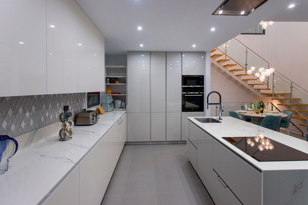 Fotografía de arquitectura interior mostrando una cocina blanca pulcra con isla, conectada estéticamente con una moderna escalera flotante