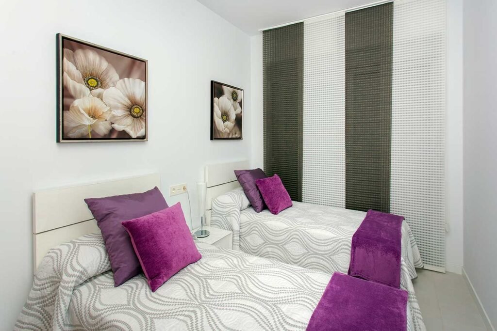 Interiorismo de habitación doble con dos camas individuales decoradas con cojines morados y cabeceros grises modernos