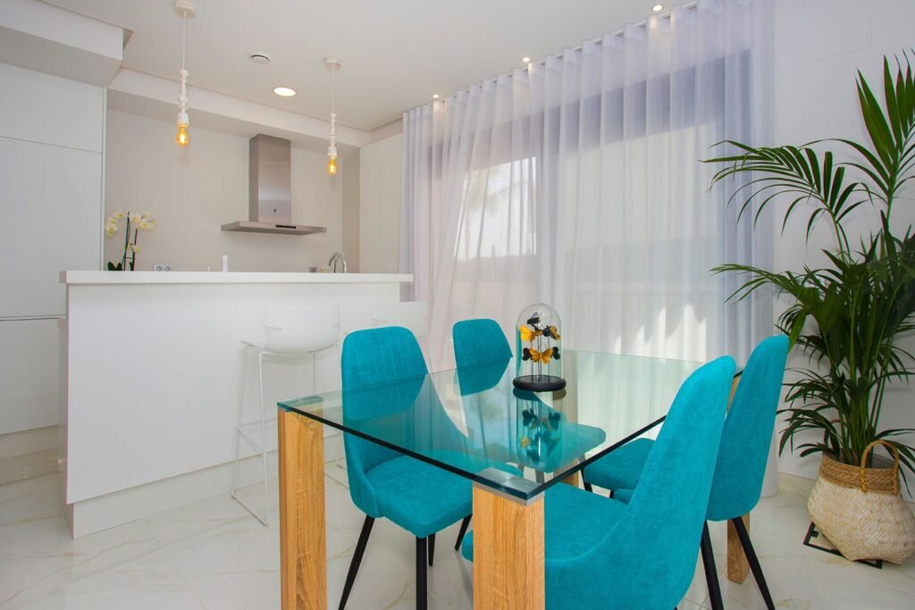 Perspectiva de salón moderno con mesa de cristal y sillas turquesas, conectado con una cocina blanca y luminosa