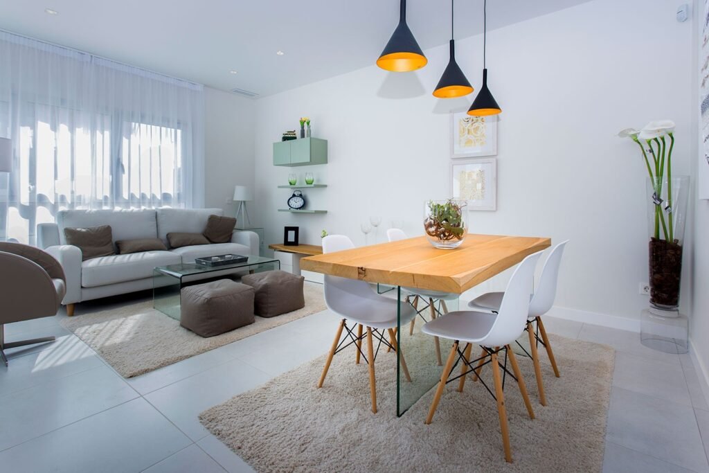 Salón comedor en tonos blancos con tres lámparas colgantes negras sobre la mesa de madera y alfombra geométrica