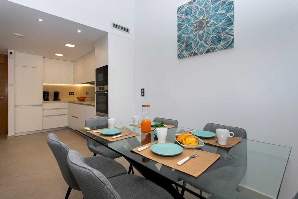 Comedor moderno con mesa de cristal, sillas de diseño tapizadas y salón al fondo con gran iluminación natural