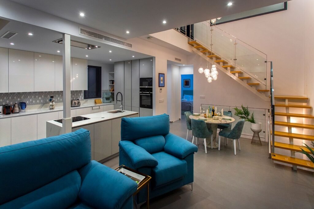 Interiorismo de salón moderno con sofá esquinero azul, mesa de centro y escalera flotante con barandilla de cristal