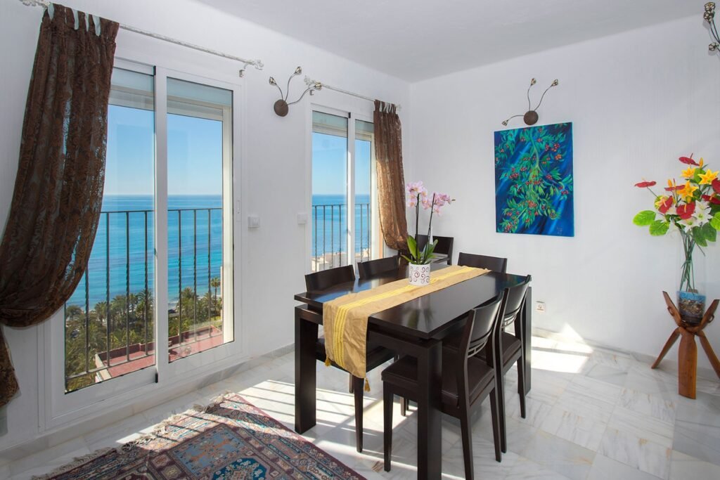 Interiorismo de salón moderno con ventanales orientados al mar, mesa de comedor larga y mucha luz natural