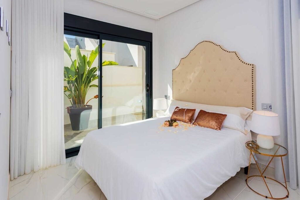 Fotografía profesional de un dormitorio minimalista con paredes blancas, cama de matrimonio impecable y detalles decorativos dorados
