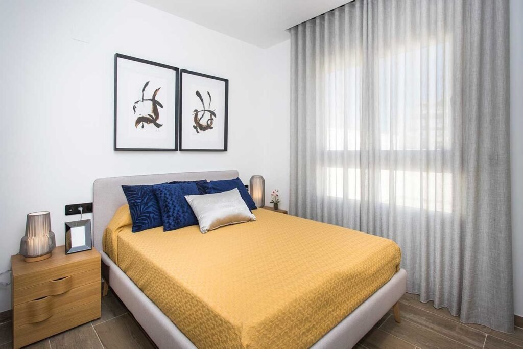 Fotografía profesional de un dormitorio de matrimonio moderno decorado con colcha mostaza, cojines azul marino, mesitas de noche de madera y cuadros minimalistas en la pared