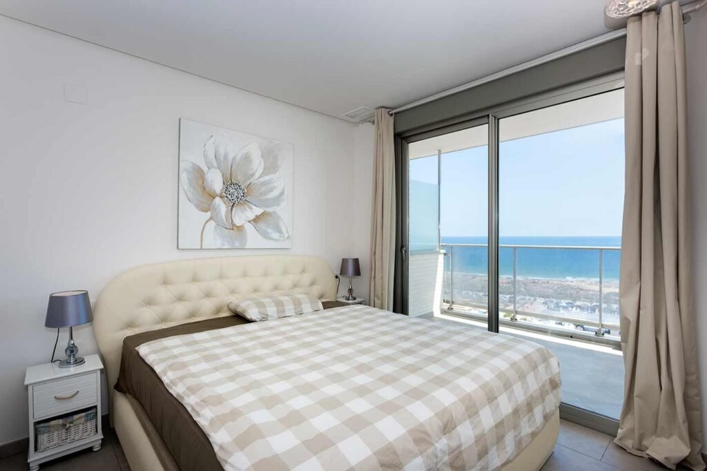 Dormitorio con cama matrimonial junto a gran ventanal corredero con vistas al mar